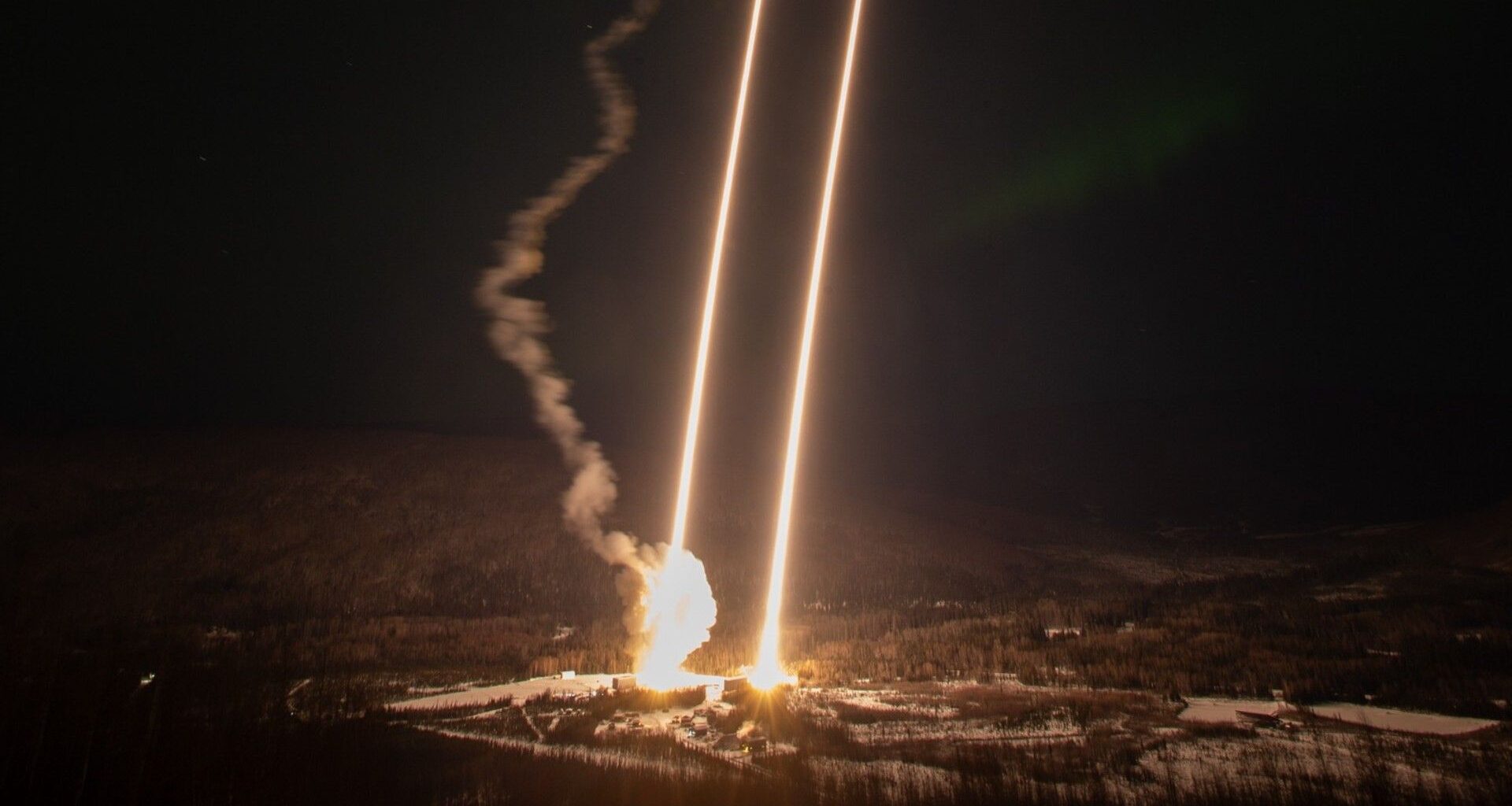 streaks of bright light reach from the ground to the sky above a snowy landscape dotted with evergreen trees