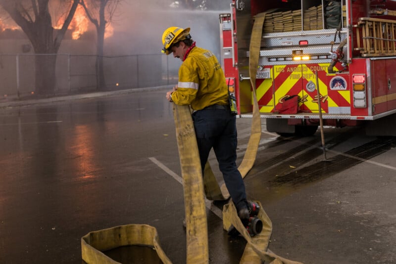 A firefighter in yellow gear unrolls a hose from a fire truck as smoke and flames rise from a building in the background. The scene appears tense and urgent, with water on the ground.