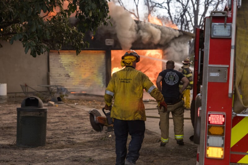 Firefighters in protective gear approach a burning building with thick smoke and flames pouring from the roof, while one firefighter holds a chainsaw near a fire truck.