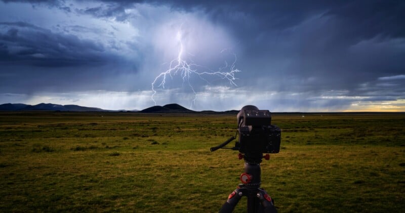 A camera on a tripod is set up in a grassy field, aimed at distant hills under a dark, stormy sky with a bolt of lightning striking in the background.