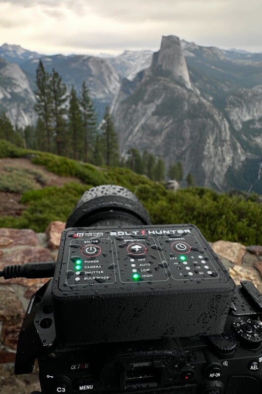 A camera with a raindrop-covered Bolt Hunter control panel is set up on a mountain overlook, facing the distant Half Dome rock formation in Yosemite National Park under a cloudy sky.
