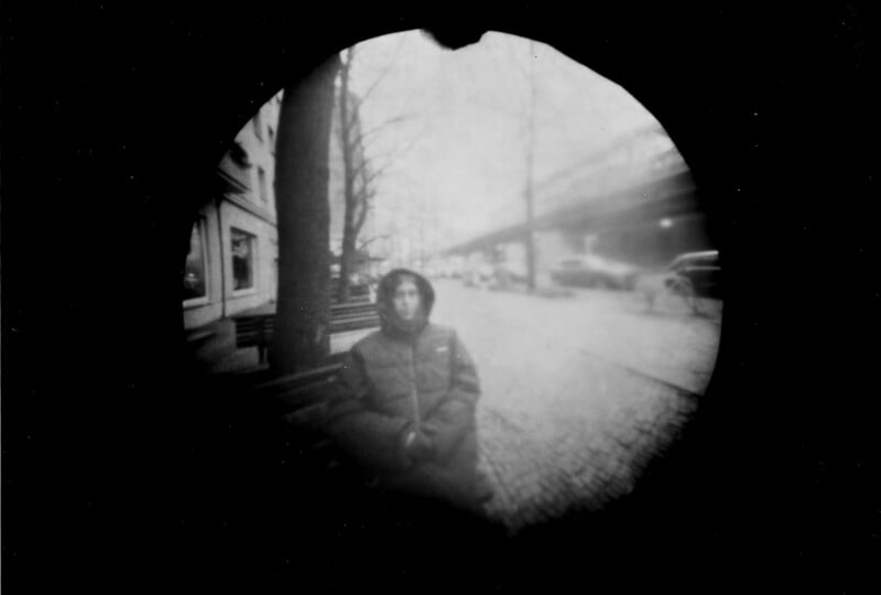 A black and white, circular, blurred photo shows a person in a hooded coat sitting on a bench by a tree along a city street with parked cars and buildings in the background.
