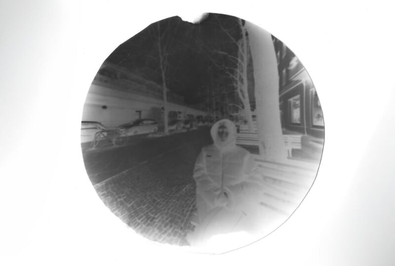 A circular black-and-white negative image shows a person sitting on a bench along a cobblestone street with bare trees, parked cars, and buildings in the background. The person's features are indistinct due to the negative effect.