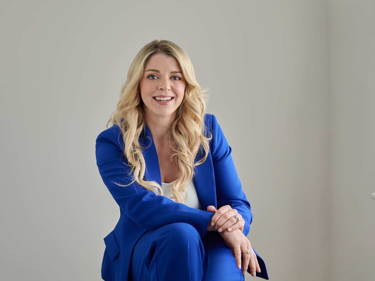 A young white blond woman in a blue suit and white shirt against a grey background.