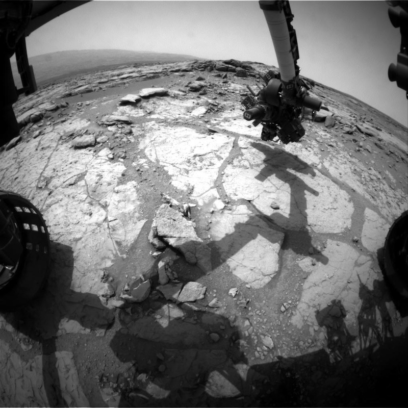 Black and white image of a group of flat whitish rocks with a robotic arm from a rover deployed over them. The rover's shadow can also be seen.