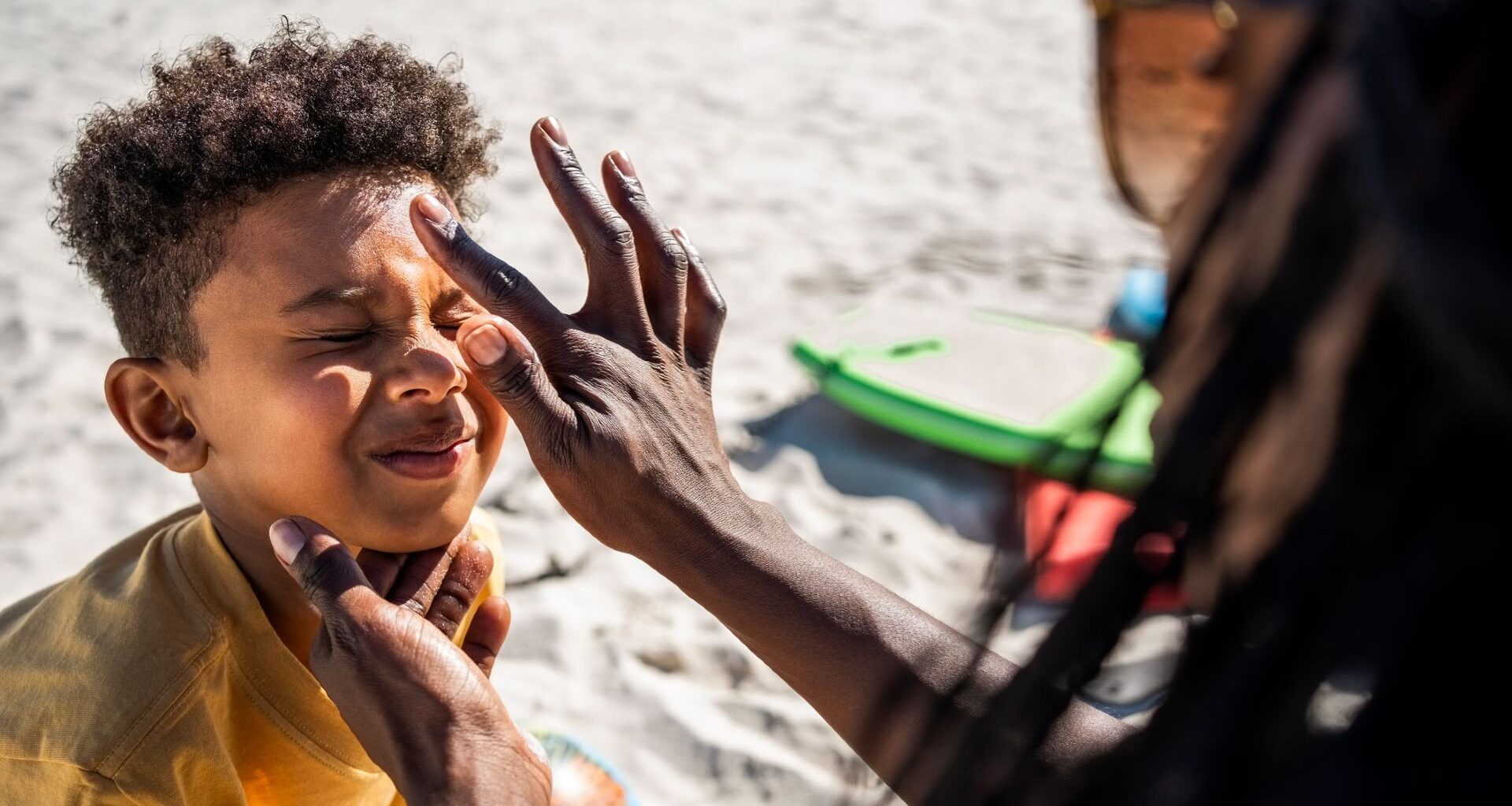 A woman wearing sunglasses with long hair applies a dot of sunscreen to a young boy who scrunches up his face in response