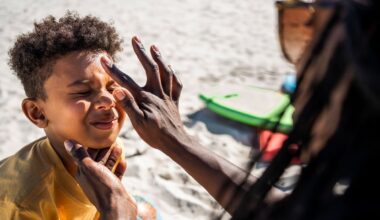 A woman wearing sunglasses with long hair applies a dot of sunscreen to a young boy who scrunches up his face in response