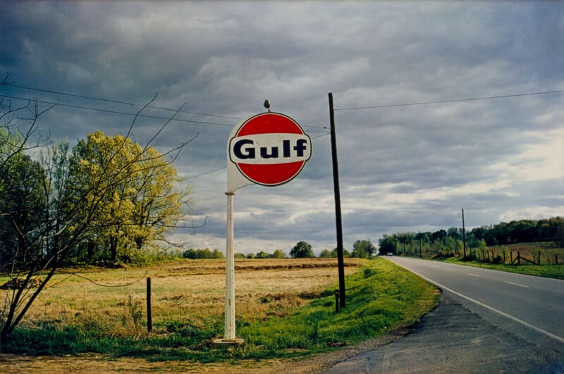 A vintage Gulf gas station sign stands beside an empty rural road with fields and trees under a cloudy sky, evoking a quiet, nostalgic countryside scene.