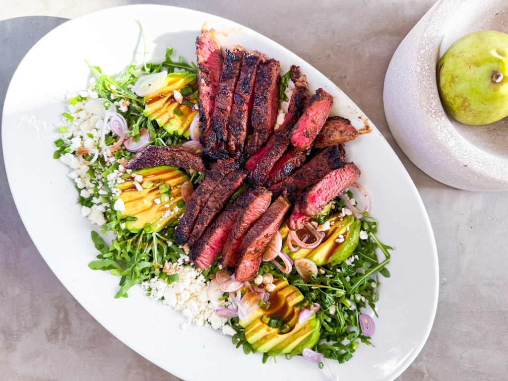 A large white plate filled with grilled steak, arugula and sliced avocado