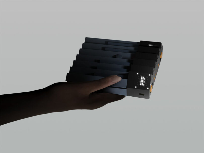 A hand holds a rectangular electronic device with a segmented design, buttons, and a small LED display showing the letter "E.