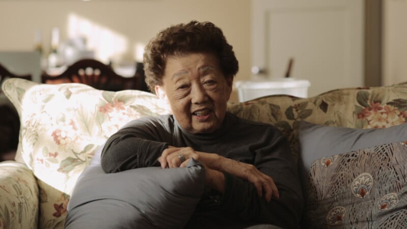An elderly woman with short curly hair sits on a floral couch, smiling warmly. She is wearing a dark long-sleeve top and is resting her arms on a blue cushion in a cozy, sunlit living room.