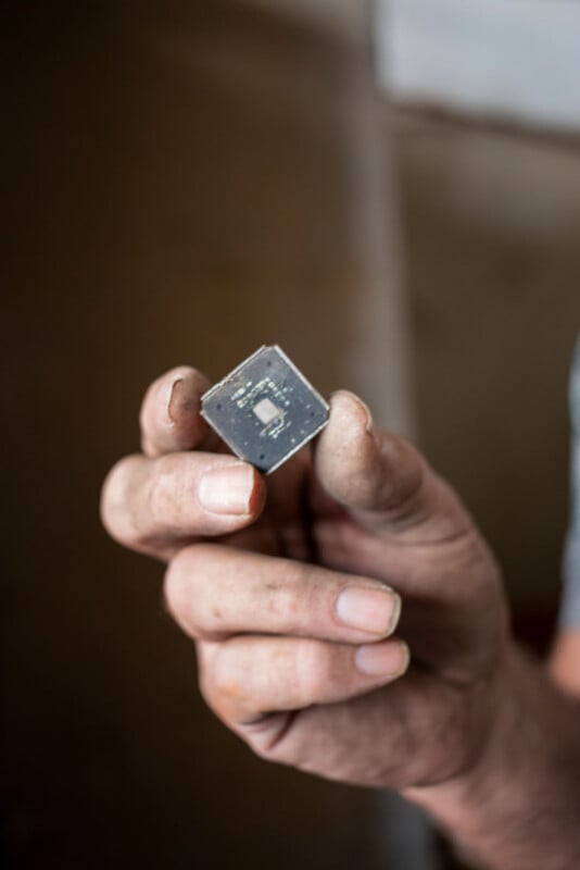A close-up of a person's dirty hand holding a small, square microchip between their fingers. The background is blurred, drawing focus to the electronic component.