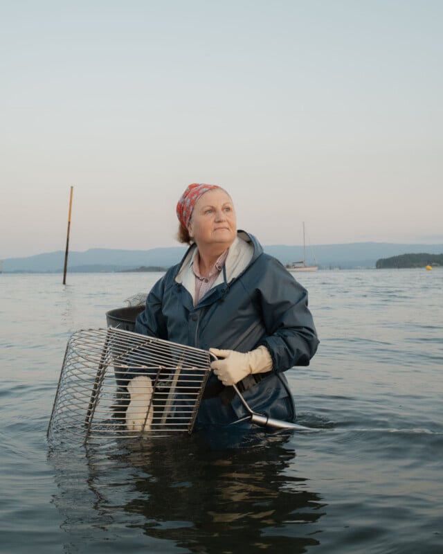 A woman in a blue raincoat and red bandana stands waist-deep in water, holding a wire crab trap. She looks off into the distance, with boats and mountains visible in the background under a clear sky.
