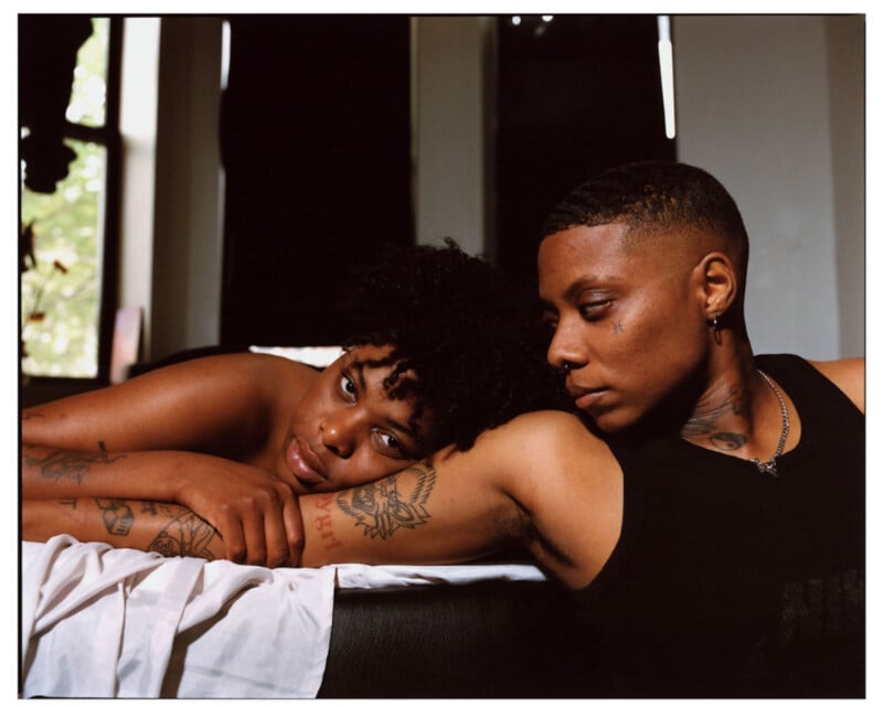 Two people with tattoos rest closely together on a bed, one lying down with their head on their arms, looking at the camera, while the other leans over, gazing softly at them. Sunlight filters through windows in the background.