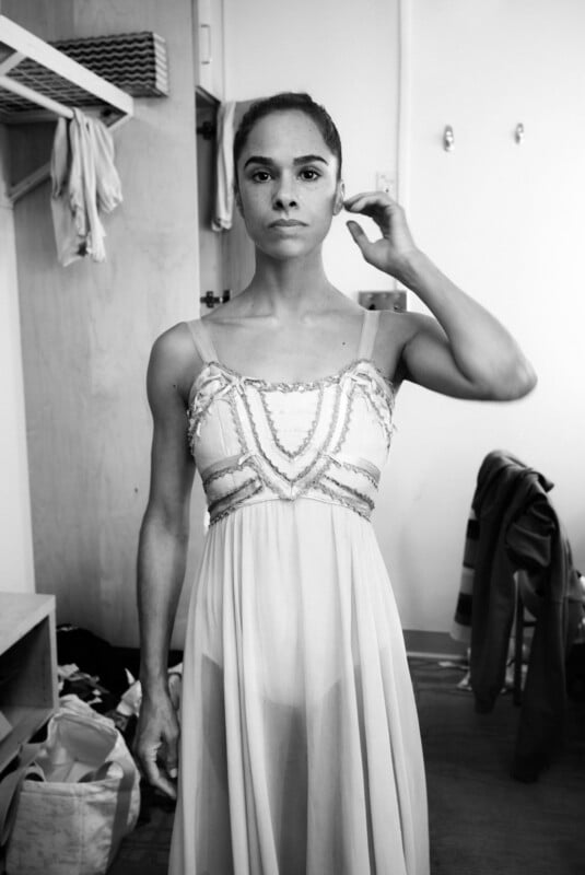 A woman in a sleeveless ballet costume stands in a dressing room, looking directly at the camera. She adjusts her hair with one hand. Clothes and bags are visible in the background. The photo is in black and white.