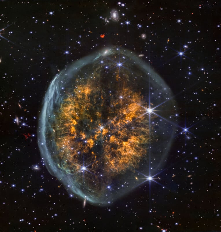 A colorful nebula in deep space, with a glowing, web-like orange and green center surrounded by a faint blue halo, against a backdrop of bright stars and distant galaxies.