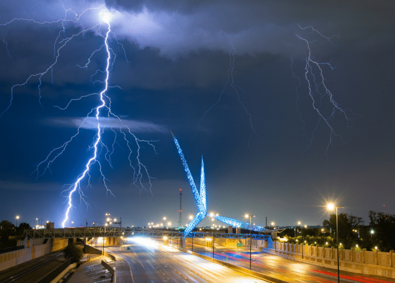 A dramatic lightning bolt strikes during a nighttime storm, illuminating the sky above a modern blue bridge over a highway with streaks of car lights, creating a vibrant and electric scene.