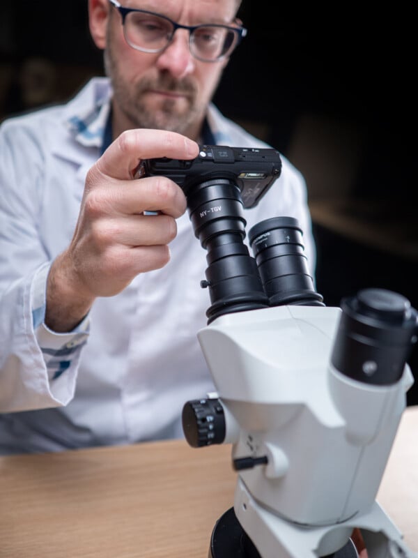 A person in a lab coat adjusts a camera attached to a microscope, focusing carefully while sitting at a desk. The background is blurred.