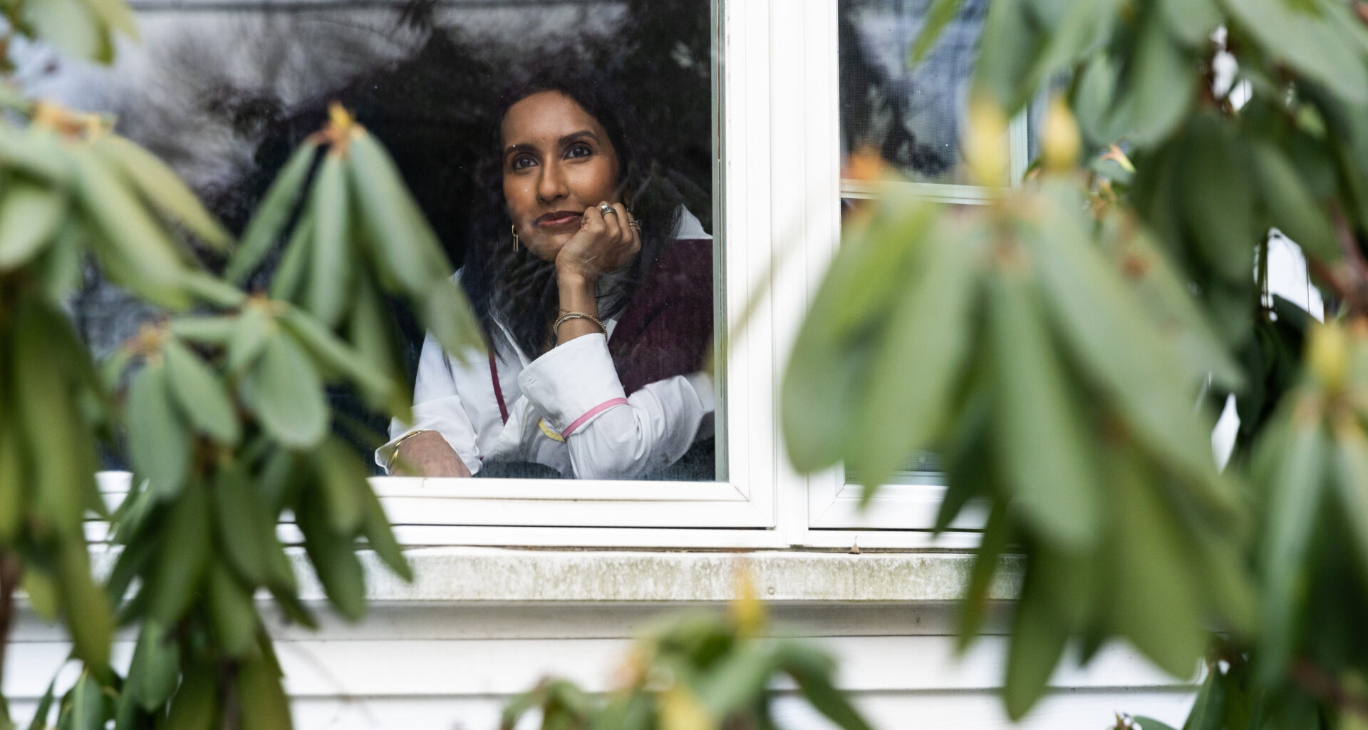 A woman looks out her window. Blurred greenery is seen in the foreground.
