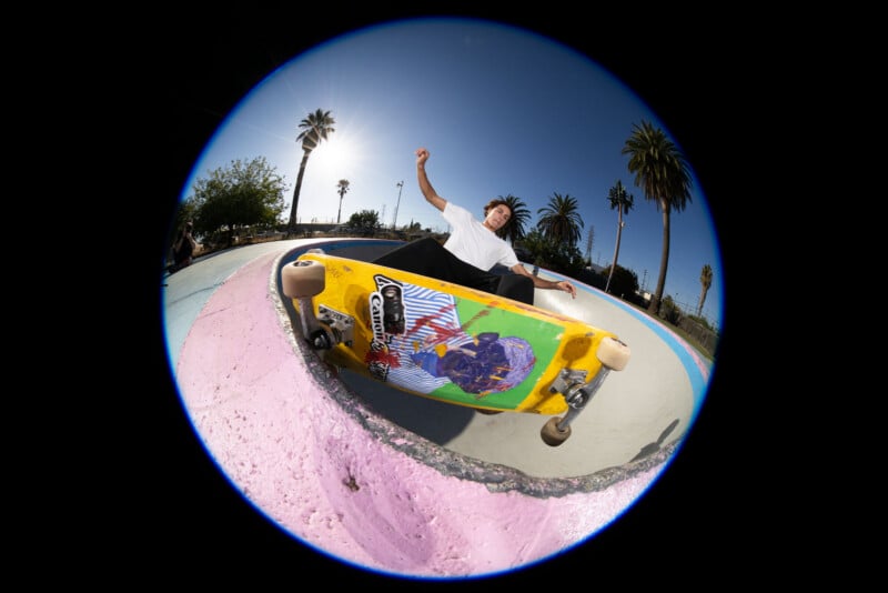 A skateboarder performs a trick on the edge of a skate bowl, captured with a fisheye lens. The bright sunlight and palm trees are visible in the background, adding a vibrant outdoor atmosphere.