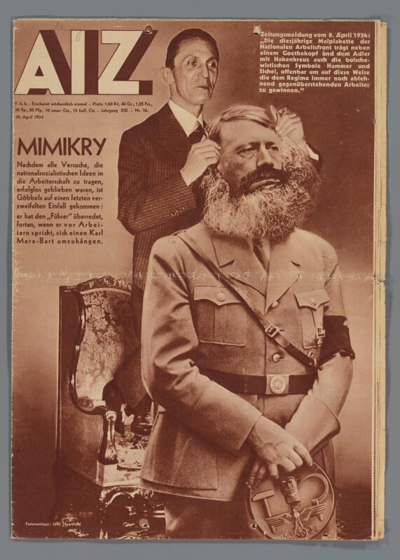 A man in a suit places a fake Karl Marx-style beard and wig onto another man wearing a Nazi uniform, with a swastika armband, in front of a mirror. The scene is sepia-toned and styled as a satirical magazine cover.
