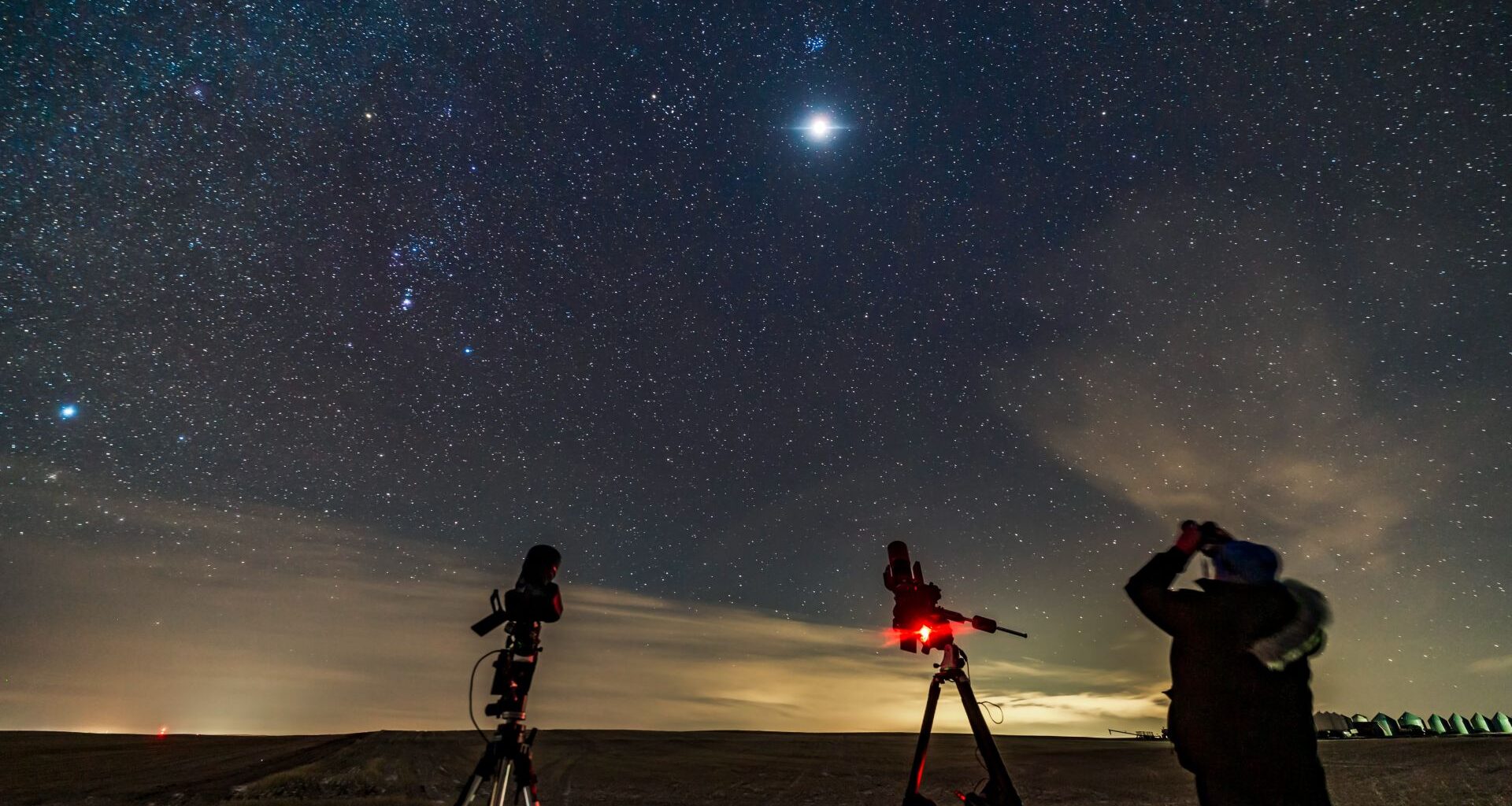 A man looks up at the moon glowing in a starry sky in a field next to two telescopes. A glow lights up clouds near the horizon.