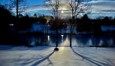 Full Snow Moon: The moon, ringed by clouds tinged with orange, among the bare branches of trees. A snowy field runs down to a creek.
