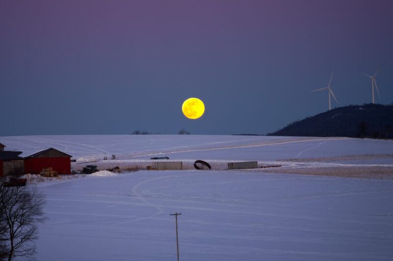 Yellow moon near the horizon, past a road, snowy fields and low hills with windmills.