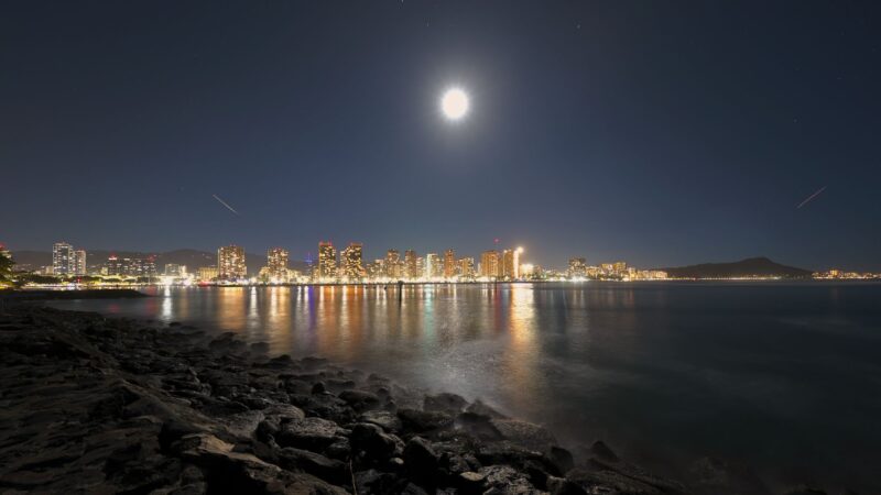 The moon over a city with its lights reflected on a calm bay. There are rocks and sand in the foreground.