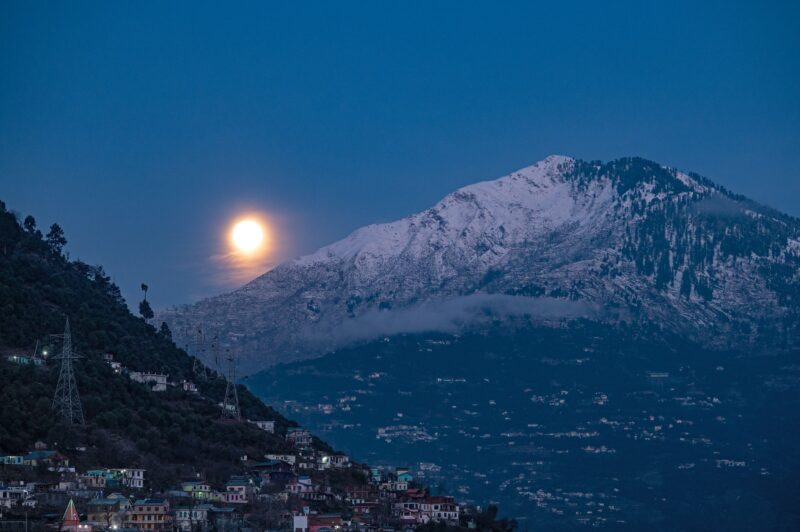 The moon shines bright next to a big mountain covered in snow, above the dim lights of a large village.