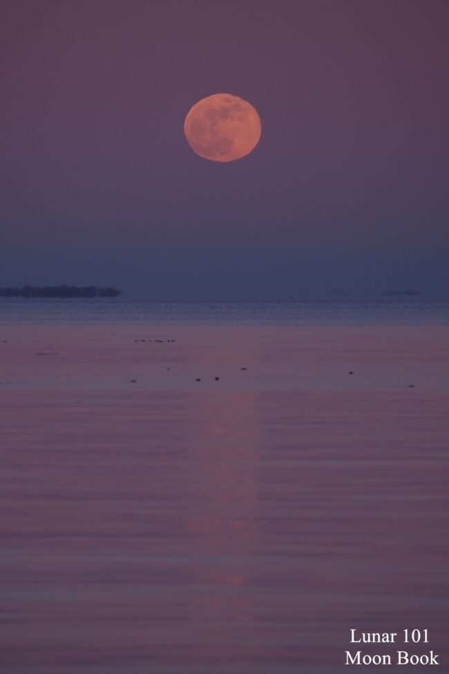 Orangish moon in a dark pink sky over water. The water below looks dark pink to blue.