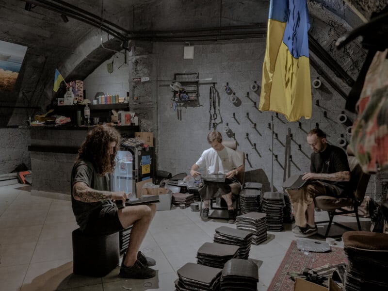 Three people sit in a room assembling items, possibly protective gear, with stacked materials around them. Ukrainian flags hang on the wall, and shelves with various supplies are in the background. The setting appears industrial.