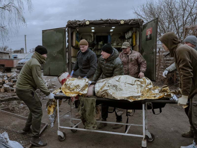 A group of people in winter clothing transports an injured person covered in a gold thermal blanket on a stretcher. They are outside, near an open military medical vehicle, on a cloudy day.