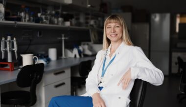 A young white woman with long blond hair in a white lab coat and blue pants poses in a biochemistry lab.