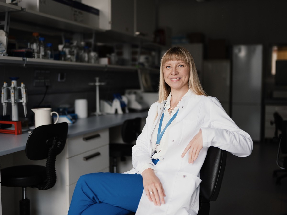 A young white woman with long blond hair in a white lab coat and blue pants poses in a biochemistry lab.
