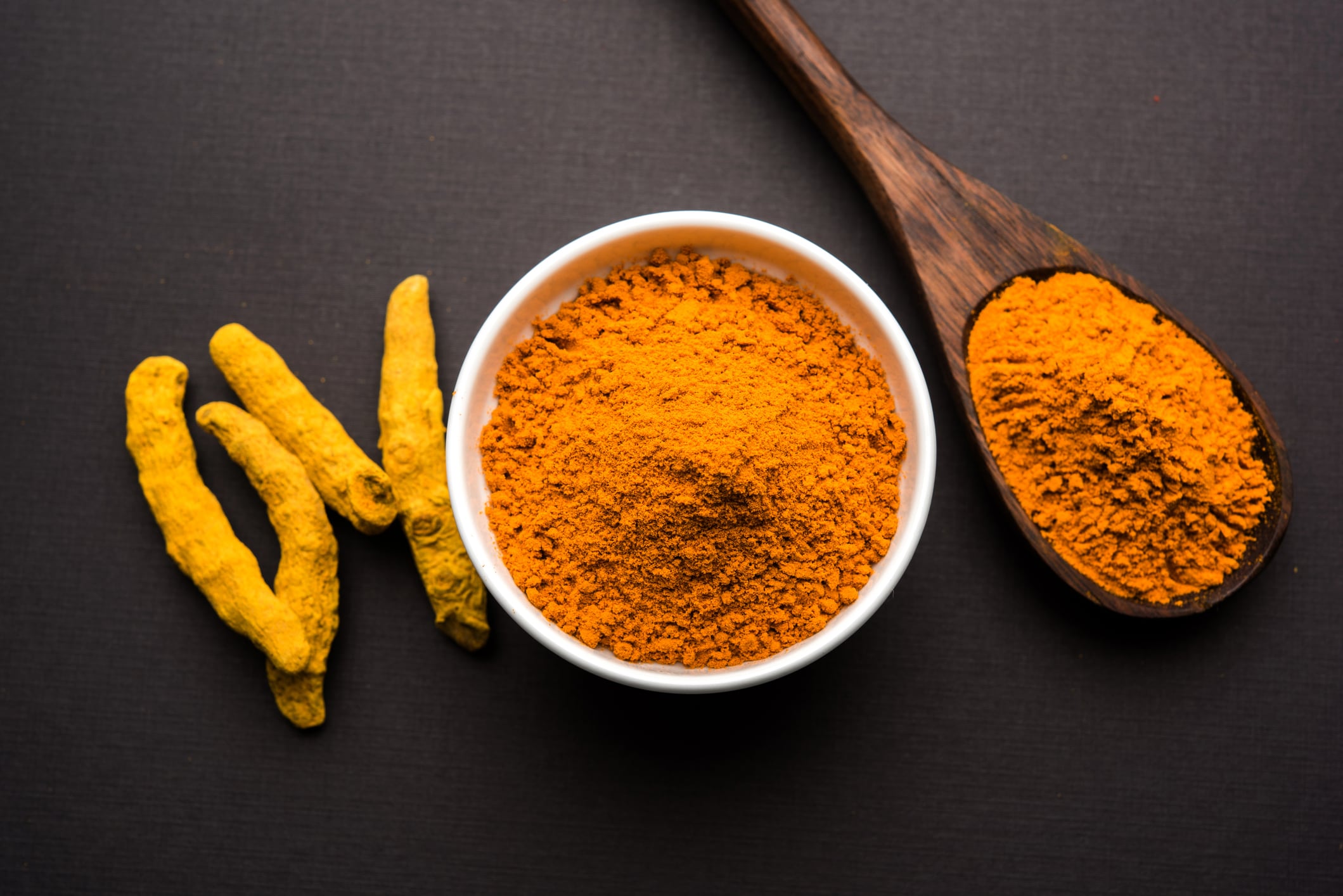 Turmeric powder in ceramic bowl with raw dried turmeric over plain background