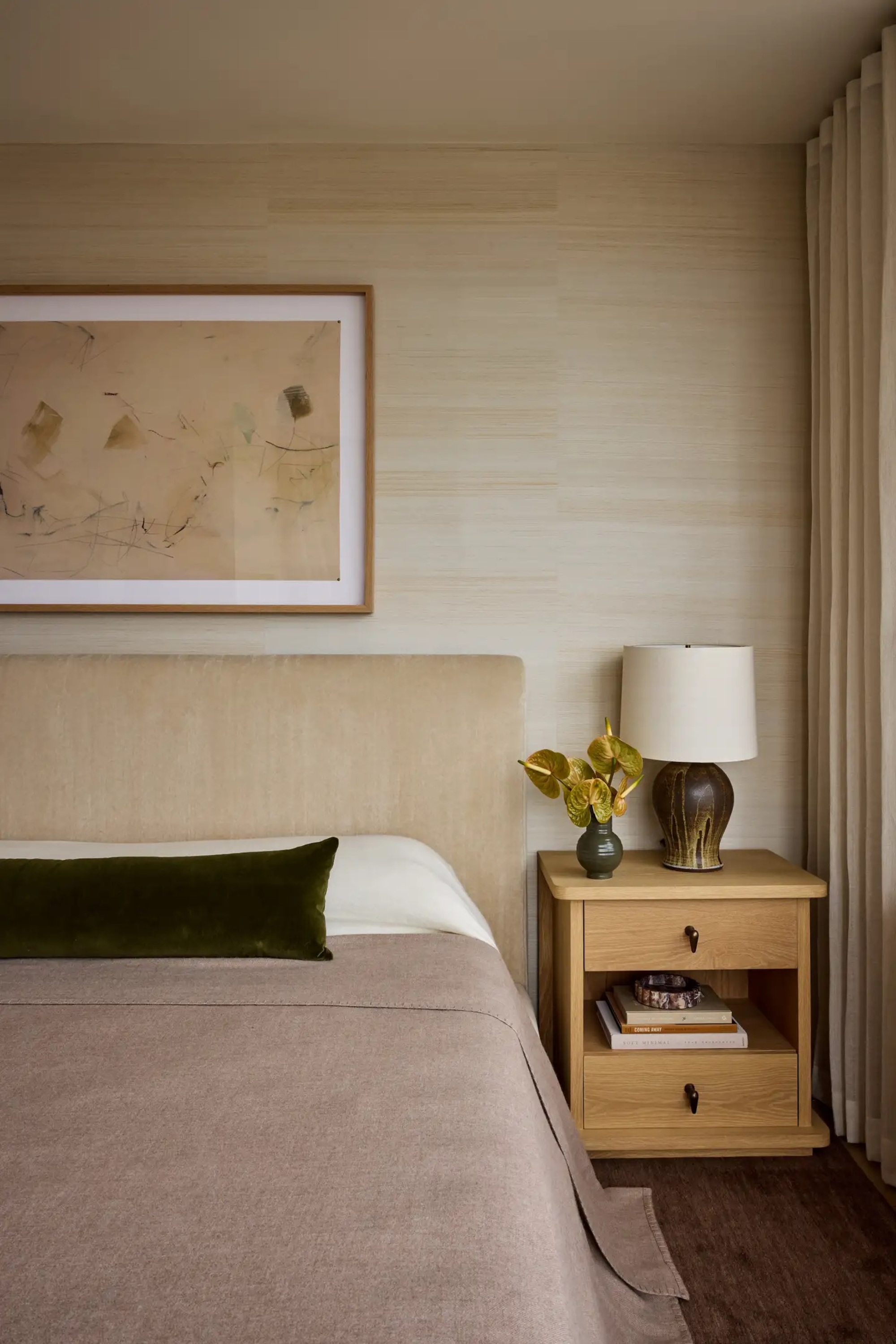 taupe bedcover with white sheets and green velvet bolster cushion, with timber side table with vase of flowers and lamp, with textured wallpaper with large artwork