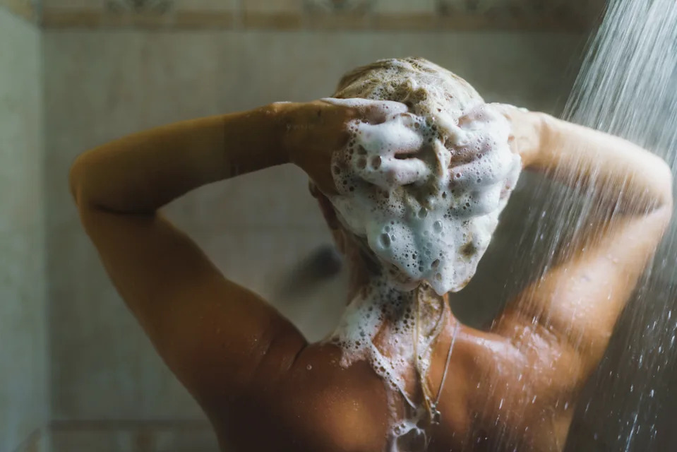 A person washing their hair in a shower, lathering shampoo with both hands