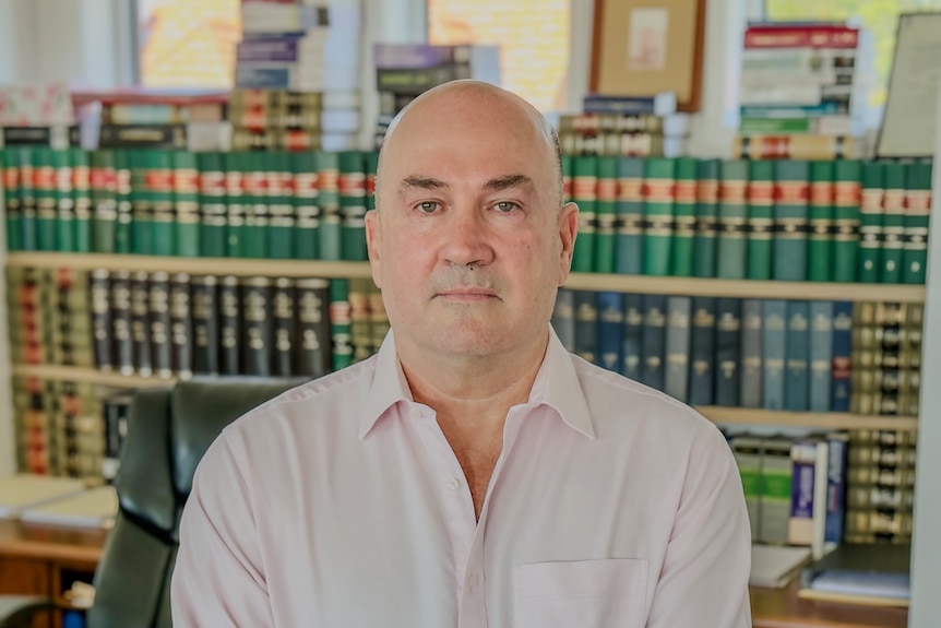 bald middle aged man in pink shirt looking at camera, bookshelf behind him