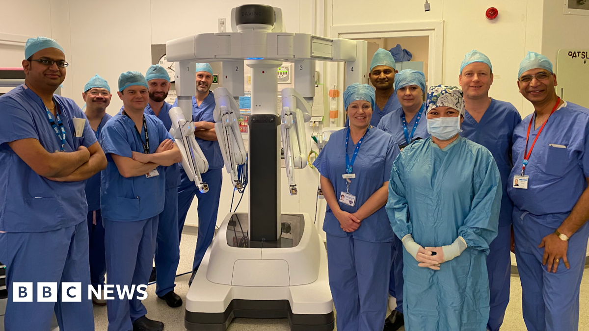A group of people dressed in blue hospital scrubs, standing around a white piece of robotic equipment. They are standing in a light-coloured room, which appears to be a hospital setting.
