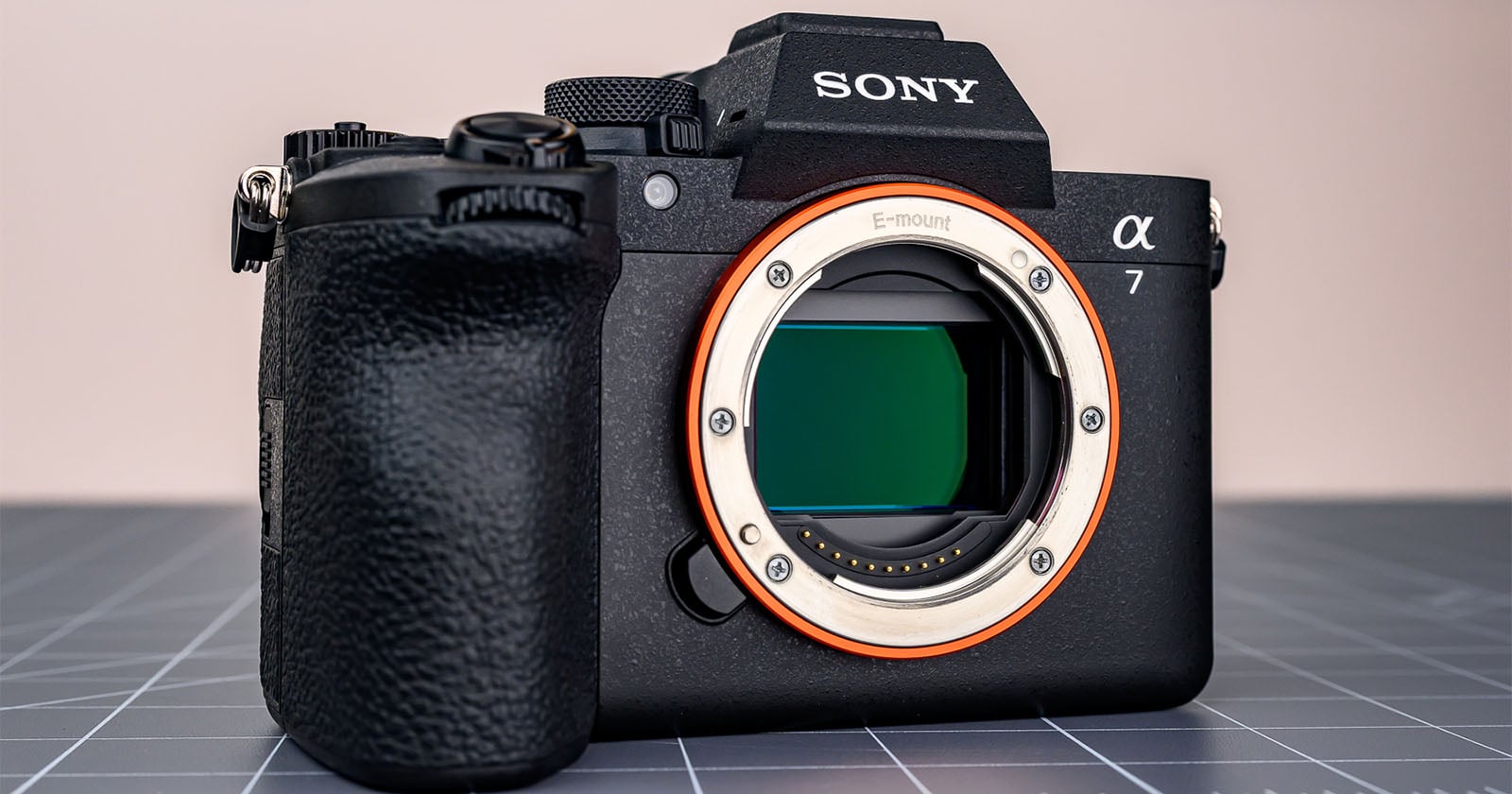 A Sony Alpha 7 mirrorless digital camera body sits on a gray grid-patterned surface, with its lens removed to reveal the camera sensor and E-mount.