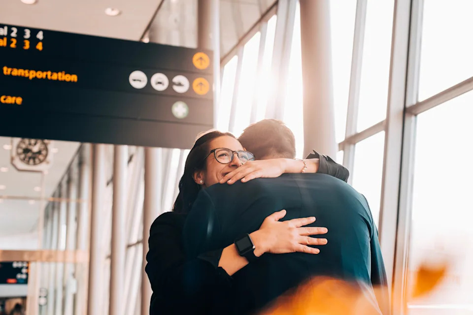 Two people embracing at an airport terminal, with signs for transportation visible