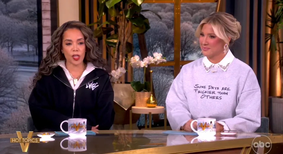 Two women sit at a talk show table wearing casual sweatshirts, engaging in conversation