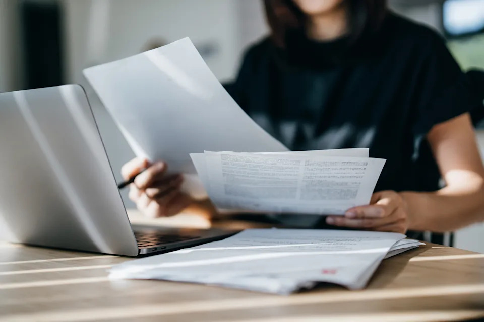 Person reviewing documents at a desk, holding papers near an open laptop. Ideal for discussions on work, productivity, or business tasks