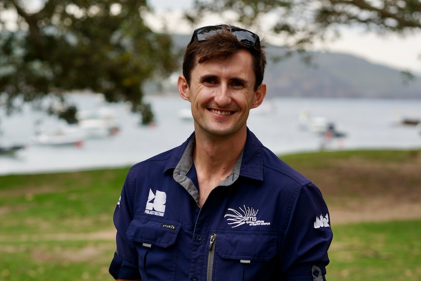 Tom Burd stands in front of a harbour park, wearing a dark jacket and glasses on his head.