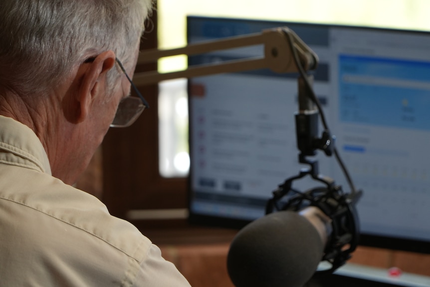 Older man in white shirt sits at radio broadcasting desk