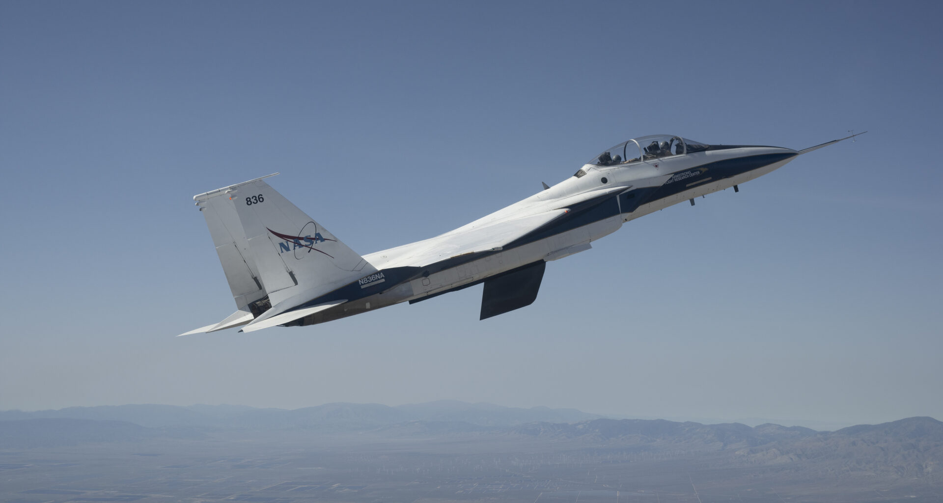 A white and blue NASA F-15 research jet climbs to altitude with an approximately 3-foot experimental wing design mounted beneath its fuselage. Viewed in profile against a blue sky with mountains in the distance, the test article resembles a ventral fin below the aircraft.