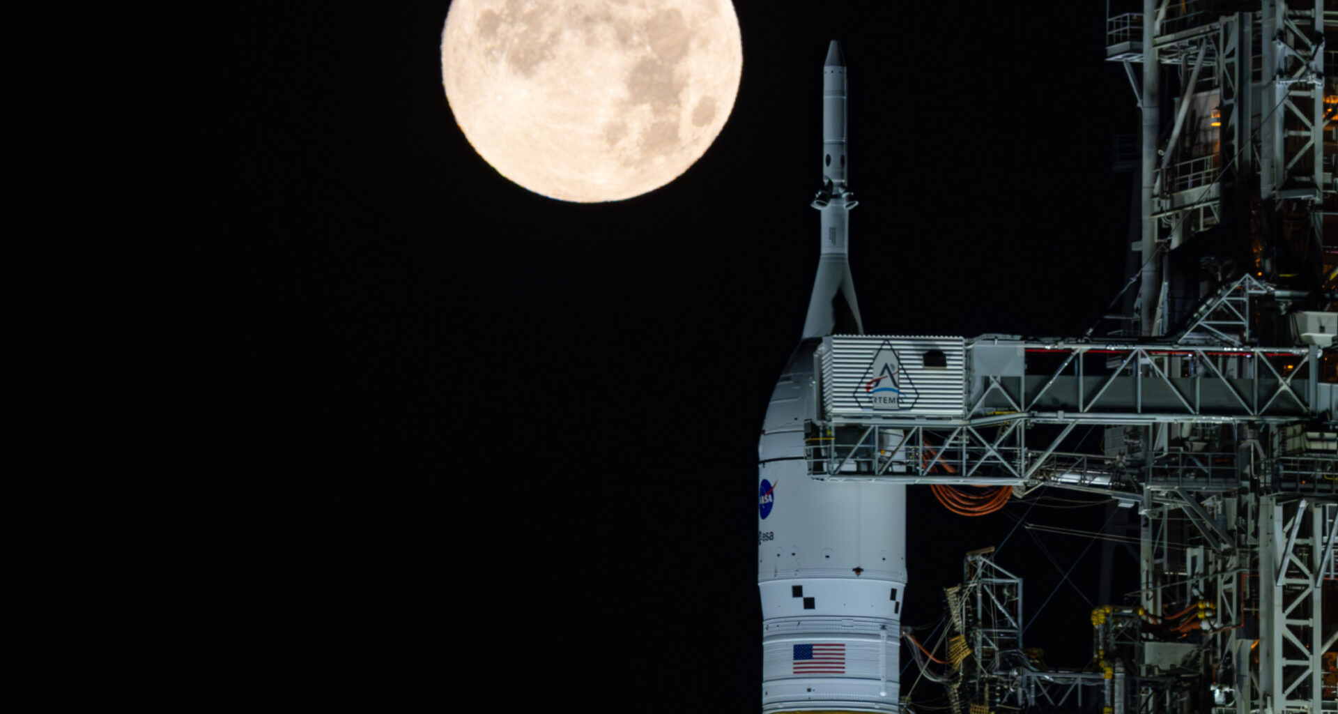 A golden full moon is seen shining over NASA’s SLS (Space Launch System) and Orion spacecraft, atop the mobile launcher in the early hours of February 1, 2026. The SLS core stage is a distinctive deep orange, while Orion is white and sits on top of the core stage.
