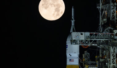A golden full moon is seen shining over NASA’s SLS (Space Launch System) and Orion spacecraft, atop the mobile launcher in the early hours of February 1, 2026. The SLS core stage is a distinctive deep orange, while Orion is white and sits on top of the core stage.