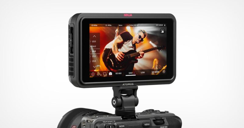 A camera monitor displays a video of a guitarist performing energetically on stage. The monitor is mounted on top of a camera, and various control icons and settings are visible on the screen.